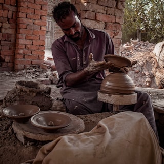 A craftsman is engaged in pottery making, shaping clay on a spinning wheel. He is surrounded by brick walls and natural lighting, with pottery pieces placed around him. His clothing appears worn with clay stains, indicating hands-on work.