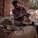 A craftsman is engaged in pottery making, shaping clay on a spinning wheel. He is surrounded by brick walls and natural lighting, with pottery pieces placed around him. His clothing appears worn with clay stains, indicating hands-on work.