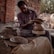 A craftsman is engaged in pottery making, shaping clay on a spinning wheel. He is surrounded by brick walls and natural lighting, with pottery pieces placed around him. His clothing appears worn with clay stains, indicating hands-on work.