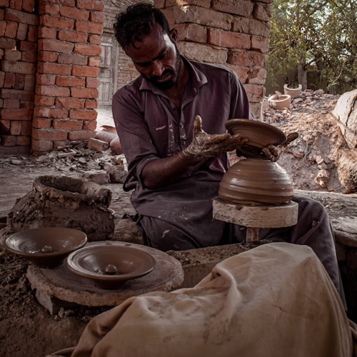A craftsman is engaged in pottery making, shaping clay on a spinning wheel. He is surrounded by brick walls and natural lighting, with pottery pieces placed around him. His clothing appears worn with clay stains, indicating hands-on work.