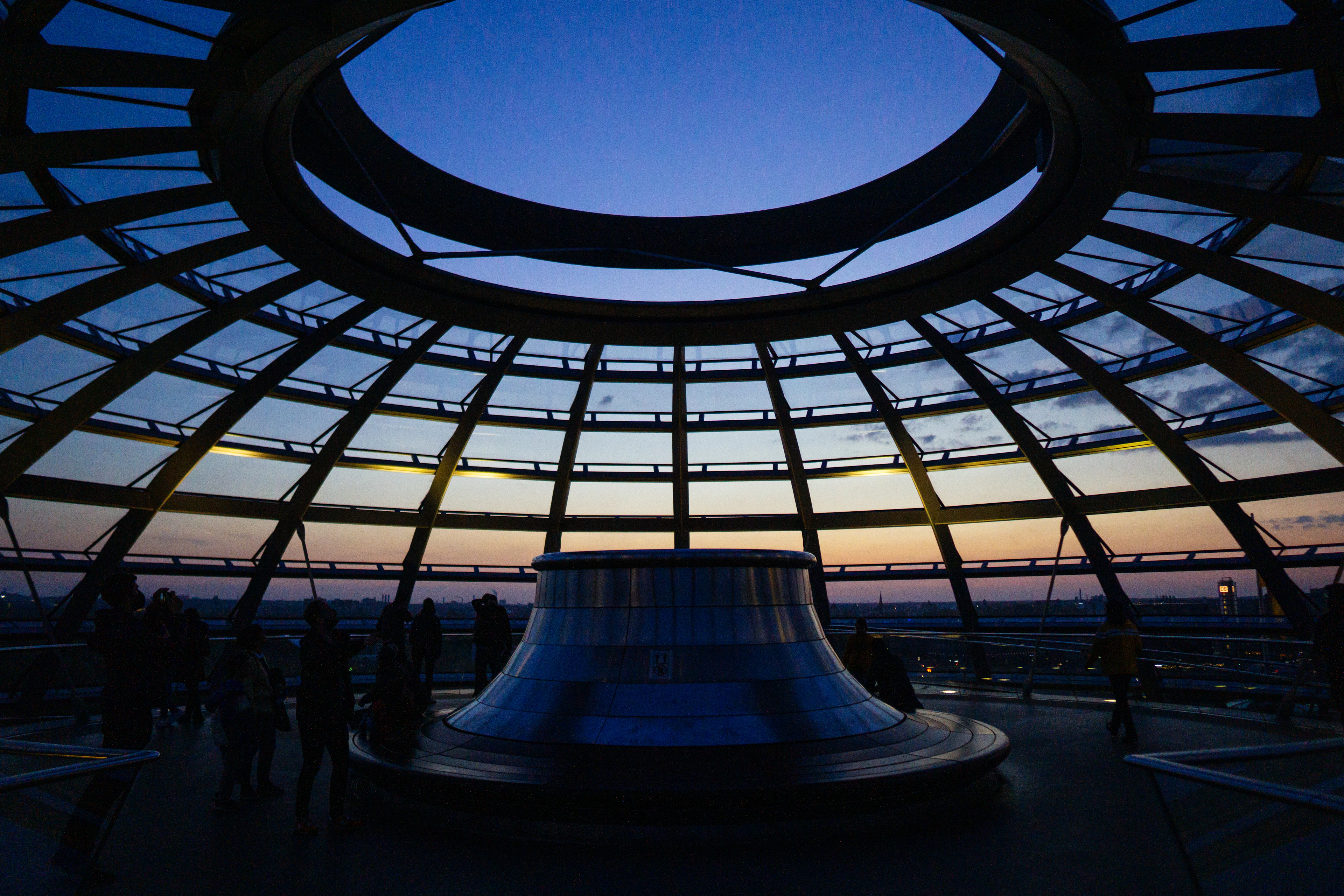 Inside the glass dome of the Reichstag at dusk, the mirrored central cone silhouetted against a sky fading from deep blue to orange through the ribbed glass, visitors small around its base.