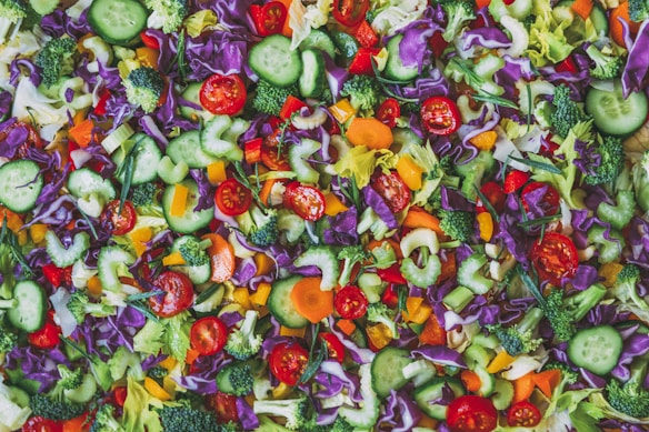 A vibrant, colorful mix of freshly chopped vegetables including sliced cucumbers, cherry tomatoes, broccoli florets, shredded purple cabbage, bell peppers, carrots, and celery arranged in a scattered pattern. The vegetables are crisp and fresh, providing a visually appealing and healthy assortment of food.