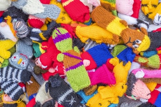 A pile of colorful knitted figures, each with distinct patterns and designs. Various characters are visible, including animals and humanoid figures, featuring bright and vibrant yarns in a mix of colors.