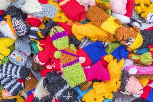A pile of colorful knitted figures, each with distinct patterns and designs. Various characters are visible, including animals and humanoid figures, featuring bright and vibrant yarns in a mix of colors.