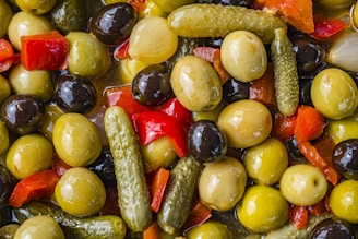 A close-up view of a mix of pickled vegetables, including green and black olives, small pickles, red bell peppers, and onions, all glistening in a liquid brine.