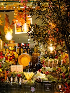 A vivid and festive display featuring a variety of wines, spirits, and gourmet foods surrounded by decorative pine branches and berries. Warm ambient lighting with hanging light bulbs and autumn foliage enhances the cozy atmosphere. A wooden cask, bottles of red wine, and a large jar filled with colorful fruits are prominently arranged.