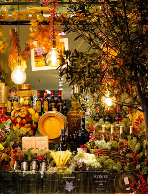 A vivid and festive display featuring a variety of wines, spirits, and gourmet foods surrounded by decorative pine branches and berries. Warm ambient lighting with hanging light bulbs and autumn foliage enhances the cozy atmosphere. A wooden cask, bottles of red wine, and a large jar filled with colorful fruits are prominently arranged.