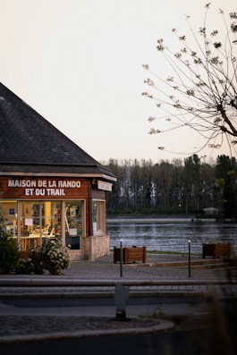 A building with large glass windows is situated near a tranquil body of water. The sign on the building reads 'Maison de la Rando et du Trail'. Trees are visible in the background against a clear sky. A tree with sparse branches and leaves is in the foreground.