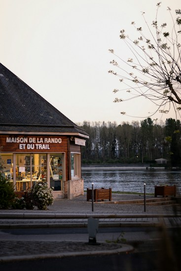 A building with large glass windows is situated near a tranquil body of water. The sign on the building reads 'Maison de la Rando et du Trail'. Trees are visible in the background against a clear sky. A tree with sparse branches and leaves is in the foreground.