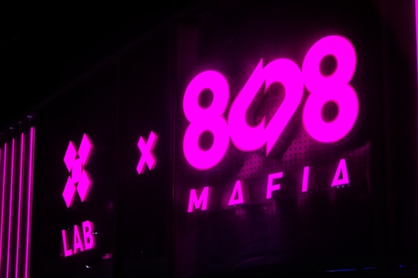 Bright pink neon signage with bold text displaying '808 Mafia' and 'LAB'. Reflective surfaces amplify the glow, creating a vibrant and urban atmosphere, typical of a nightclub or music studio.