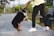 A person wearing a yellow jacket and black pants is playing with a tricolor dog on a paved pathway. The dog is standing on its hind legs, reaching for a yellow and black toy held by the person. In the background, there are green trees and a park bench.