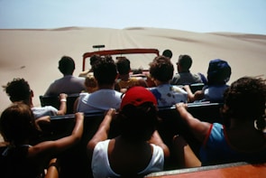 A group of people are seated in an open-top vehicle driving through a vast desert landscape with sand dunes visible in the distance. Most of the individuals have their backs turned, suggesting they are focused on the journey ahead.