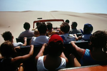A group of people are seated in an open-top vehicle driving through a vast desert landscape with sand dunes visible in the distance. Most of the individuals have their backs turned, suggesting they are focused on the journey ahead.