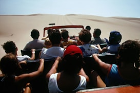 A group of people are seated in an open-top vehicle driving through a vast desert landscape with sand dunes visible in the distance. Most of the individuals have their backs turned, suggesting they are focused on the journey ahead.