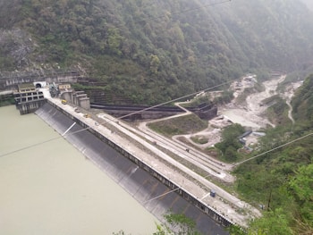 A large dam structure with a sloped concrete wall holds back a body of water. Surrounding the dam are lush green hills covered with dense vegetation. A road runs along the top of the dam with several vehicles visible. The scene is enveloped in a light mist, adding a serene quality to the landscape.