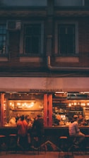 A cozy cafe scene with a group of people sitting at a bar counter inside a dimly lit bagel shop. Warm lighting from hanging bulbs casts a soft glow on the patrons, who appear to be engaged in conversation and enjoying their time. The interior features exposed brick walls and shelves filled with bottles.