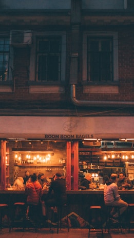 A cozy cafe scene with a group of people sitting at a bar counter inside a dimly lit bagel shop. Warm lighting from hanging bulbs casts a soft glow on the patrons, who appear to be engaged in conversation and enjoying their time. The interior features exposed brick walls and shelves filled with bottles.