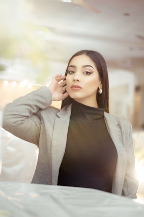 A person with long dark hair wears a gray blazer over a black turtleneck, adorned with elegant gold earrings. Sunlight creates a soft glow, enhancing the serene and sophisticated ambiance. The blurred background suggests an indoor setting, possibly a restaurant or cafe.