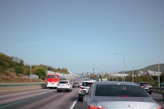 A highway scene with multiple vehicles, including cars and a red bus, traveling in the same direction. The road is bordered by green hills and trees, with several streetlights along the roadside. In the distance, there are buildings visible under a clear blue sky, indicating an urban area approaching.