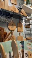 A collection of wooden kitchen utensils and cutting boards displayed on shelves. There are various sizes and shapes of cutting boards, with some featuring engraved text like 'bon appétit' and 'made with love.' A rack of knives is also visible, along with a shelf holding a plant, and some neatly arranged cooking spoons in labeled packaging.