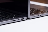 Two sleek, modern laptops positioned back-to-back on a light surface, showcasing the sleek edges and ports of the devices. The keyboards are partially visible, and the lighting highlights the metallic texture of the laptops.