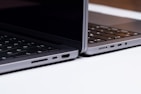Two sleek, modern laptops positioned back-to-back on a light surface, showcasing the sleek edges and ports of the devices. The keyboards are partially visible, and the lighting highlights the metallic texture of the laptops.
