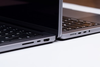 Two sleek, modern laptops positioned back-to-back on a light surface, showcasing the sleek edges and ports of the devices. The keyboards are partially visible, and the lighting highlights the metallic texture of the laptops.