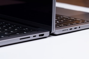 Two sleek, modern laptops positioned back-to-back on a light surface, showcasing the sleek edges and ports of the devices. The keyboards are partially visible, and the lighting highlights the metallic texture of the laptops.