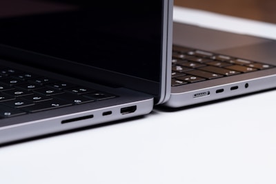 Two sleek, modern laptops positioned back-to-back on a light surface, showcasing the sleek edges and ports of the devices. The keyboards are partially visible, and the lighting highlights the metallic texture of the laptops.