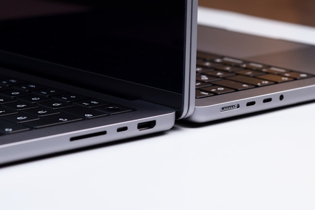 Two sleek, modern laptops positioned back-to-back on a light surface, showcasing the sleek edges and ports of the devices. The keyboards are partially visible, and the lighting highlights the metallic texture of the laptops.