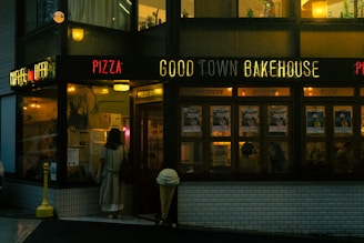 A person in a trench coat is standing outside a dimly lit bakery and restaurant named 'Good Town Bakehouse.' The establishment's front is decorated with neon signs that read 'Pizza,' 'Good Town Bakehouse,' and 'Coffee and Beer.' The interior is cozy, illuminated with warm lights, and there are several posters visible on the windows.