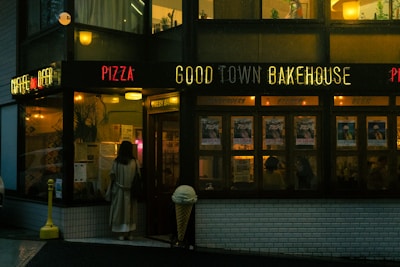 A person in a trench coat is standing outside a dimly lit bakery and restaurant named 'Good Town Bakehouse.' The establishment's front is decorated with neon signs that read 'Pizza,' 'Good Town Bakehouse,' and 'Coffee and Beer.' The interior is cozy, illuminated with warm lights, and there are several posters visible on the windows.