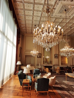 A luxurious interior with ornate chandeliers hanging from a detailed, patterned ceiling. The space is furnished with elegant chairs and tables, featuring plush upholstery in rich colors. Large windows with sheer curtains allow natural light to flood the room, accentuating the intricate design elements. A series of mirrors and framed art pieces adorn the walls, enhancing the grandeur of the setting.