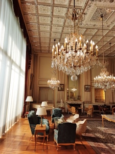 A luxurious interior with ornate chandeliers hanging from a detailed, patterned ceiling. The space is furnished with elegant chairs and tables, featuring plush upholstery in rich colors. Large windows with sheer curtains allow natural light to flood the room, accentuating the intricate design elements. A series of mirrors and framed art pieces adorn the walls, enhancing the grandeur of the setting.