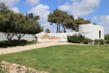 A scenic outdoor area with a well-maintained grassy lawn and a paved walkway leading to a white, circular structure. The structure features text in Hebrew and is partially surrounded by green shrubs. The foreground includes neatly trimmed hedges and trees providing shade. The sky is mostly clear with some fluffy clouds.