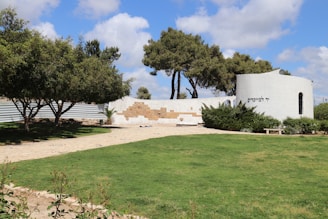 A scenic outdoor area with a well-maintained grassy lawn and a paved walkway leading to a white, circular structure. The structure features text in Hebrew and is partially surrounded by green shrubs. The foreground includes neatly trimmed hedges and trees providing shade. The sky is mostly clear with some fluffy clouds.