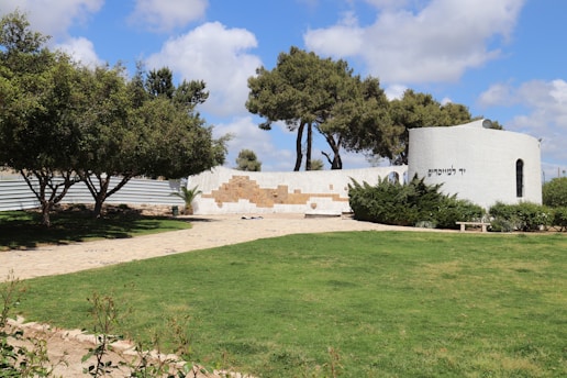 A scenic outdoor area with a well-maintained grassy lawn and a paved walkway leading to a white, circular structure. The structure features text in Hebrew and is partially surrounded by green shrubs. The foreground includes neatly trimmed hedges and trees providing shade. The sky is mostly clear with some fluffy clouds.