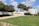 A scenic outdoor area with a well-maintained grassy lawn and a paved walkway leading to a white, circular structure. The structure features text in Hebrew and is partially surrounded by green shrubs. The foreground includes neatly trimmed hedges and trees providing shade. The sky is mostly clear with some fluffy clouds.
