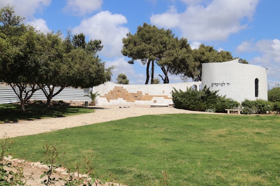 A scenic outdoor area with a well-maintained grassy lawn and a paved walkway leading to a white, circular structure. The structure features text in Hebrew and is partially surrounded by green shrubs. The foreground includes neatly trimmed hedges and trees providing shade. The sky is mostly clear with some fluffy clouds.