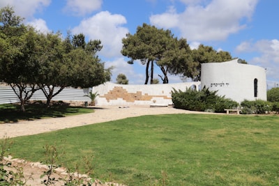 A scenic outdoor area with a well-maintained grassy lawn and a paved walkway leading to a white, circular structure. The structure features text in Hebrew and is partially surrounded by green shrubs. The foreground includes neatly trimmed hedges and trees providing shade. The sky is mostly clear with some fluffy clouds.