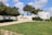 A scenic outdoor area with a well-maintained grassy lawn and a paved walkway leading to a white, circular structure. The structure features text in Hebrew and is partially surrounded by green shrubs. The foreground includes neatly trimmed hedges and trees providing shade. The sky is mostly clear with some fluffy clouds.