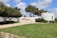 A scenic outdoor area with a well-maintained grassy lawn and a paved walkway leading to a white, circular structure. The structure features text in Hebrew and is partially surrounded by green shrubs. The foreground includes neatly trimmed hedges and trees providing shade. The sky is mostly clear with some fluffy clouds.