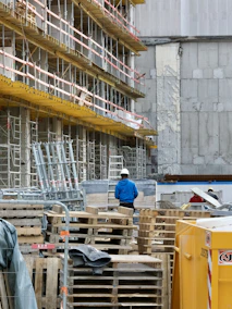 A construction site with scaffolding lining the side of a building. Numerous wooden pallets are stacked in front, along with a few metal supports. A person wearing a blue jacket and white hard hat stands among the equipment, facing away. A large yellow container is visible in the foreground. The building behind has patches of exposed concrete and weathered walls.