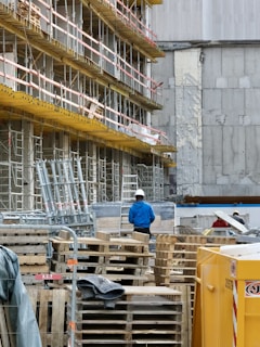 A construction site with scaffolding lining the side of a building. Numerous wooden pallets are stacked in front, along with a few metal supports. A person wearing a blue jacket and white hard hat stands among the equipment, facing away. A large yellow container is visible in the foreground. The building behind has patches of exposed concrete and weathered walls.