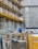 A construction site with scaffolding lining the side of a building. Numerous wooden pallets are stacked in front, along with a few metal supports. A person wearing a blue jacket and white hard hat stands among the equipment, facing away. A large yellow container is visible in the foreground. The building behind has patches of exposed concrete and weathered walls.