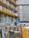 A construction site with scaffolding lining the side of a building. Numerous wooden pallets are stacked in front, along with a few metal supports. A person wearing a blue jacket and white hard hat stands among the equipment, facing away. A large yellow container is visible in the foreground. The building behind has patches of exposed concrete and weathered walls.