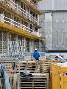 A construction site with scaffolding lining the side of a building. Numerous wooden pallets are stacked in front, along with a few metal supports. A person wearing a blue jacket and white hard hat stands among the equipment, facing away. A large yellow container is visible in the foreground. The building behind has patches of exposed concrete and weathered walls.