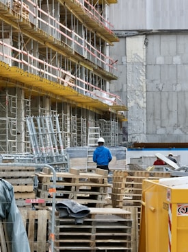 A construction site with scaffolding lining the side of a building. Numerous wooden pallets are stacked in front, along with a few metal supports. A person wearing a blue jacket and white hard hat stands among the equipment, facing away. A large yellow container is visible in the foreground. The building behind has patches of exposed concrete and weathered walls.