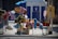 A street stand displays a variety of colorful helium balloons shaped like popular animated characters. The balloons are tied to carts, with some depicting superheroes and cartoon animals. In the background, there is a blue and white structure that resembles a portable restroom or kiosk. A few other signs and balloons can be seen, and the area is sunlit, suggesting a fair or market setting.
