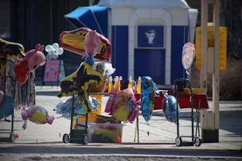 A street stand displays a variety of colorful helium balloons shaped like popular animated characters. The balloons are tied to carts, with some depicting superheroes and cartoon animals. In the background, there is a blue and white structure that resembles a portable restroom or kiosk. A few other signs and balloons can be seen, and the area is sunlit, suggesting a fair or market setting.